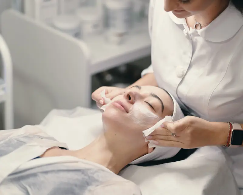 A professional wearing a white coat applies a product to a client's shoulder in a well-equipped treatment room.